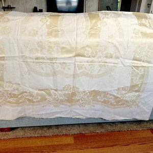 May include: Cream-colored tablecloth with a floral pattern. The tablecloth has a repeating design of flowers and leaves in a lighter cream color, creating a subtle contrast. The edges feature a decorative border.