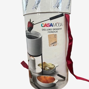 May include: A cylindrical white cardboard packaging box for a CasaModa Deluxe Dessert Fondue set. The exterior features graphics of a white fondue pot, a tea light candle, and small bowls with fruit. A red ribbon is attached to the side of the lid.