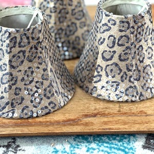 Può includere: Diverse paralumi piccoli a forma di cono con motivo leopardato. I paralumi sono beige con macchie marrone scuro e sono impreziositi da piccole paillettes argentate. Sono appoggiati su una superficie di legno.