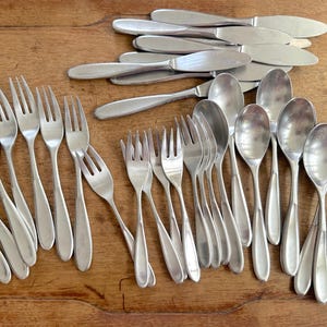 May include: A selection of stainless steel cutlery, comprising forks, spoons, and knives, displayed on a wooden surface. The silverware features a straightforward, elegant design, indicative of a classic style. The arrangement implies a dining set.