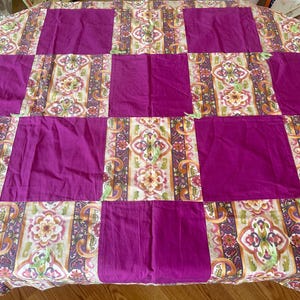 May include: A round tablecloth with a patchwork design. The design features alternating squares of solid magenta fabric and patterned fabric with floral and paisley motifs in shades of cream, green, and purple.