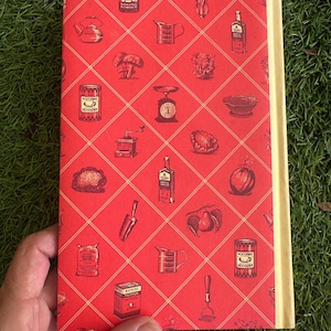 May include: A vintage cookbook with a red cover featuring a diamond pattern and illustrations of kitchen items like a teapot, scale, and spice tins. The spine is a light beige color.