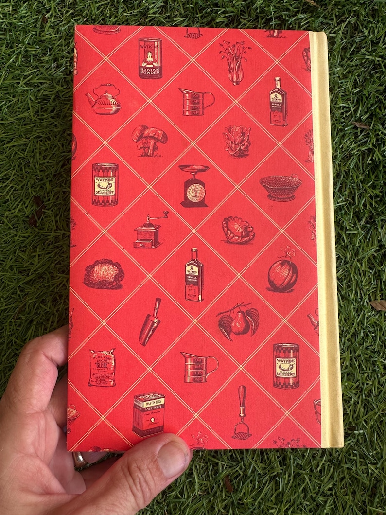 May include: A vintage cookbook with a red cover featuring a diamond pattern and illustrations of kitchen items like a teapot, scale, and spice tins. The spine is a light beige color.