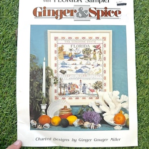 Könnte beinhalten: Ein Kreuzstichmuster mit dem Titel "FLORIDA Sampler" von Ginger & Spice. Das Design zeigt eine gerahmte Darstellung von Wahrzeichen und Szenen Floridas mit dem Text "THE SUNSHINE STATE" und "FLORIDA". Das Bild ist von dekorativen Elementen umgeben.