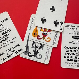 May include: A collection of vintage playing cards, including a five of clubs and two joker cards, arranged on a vibrant red surface. The cards feature classic designs and text from the Whitman Publishing Company.