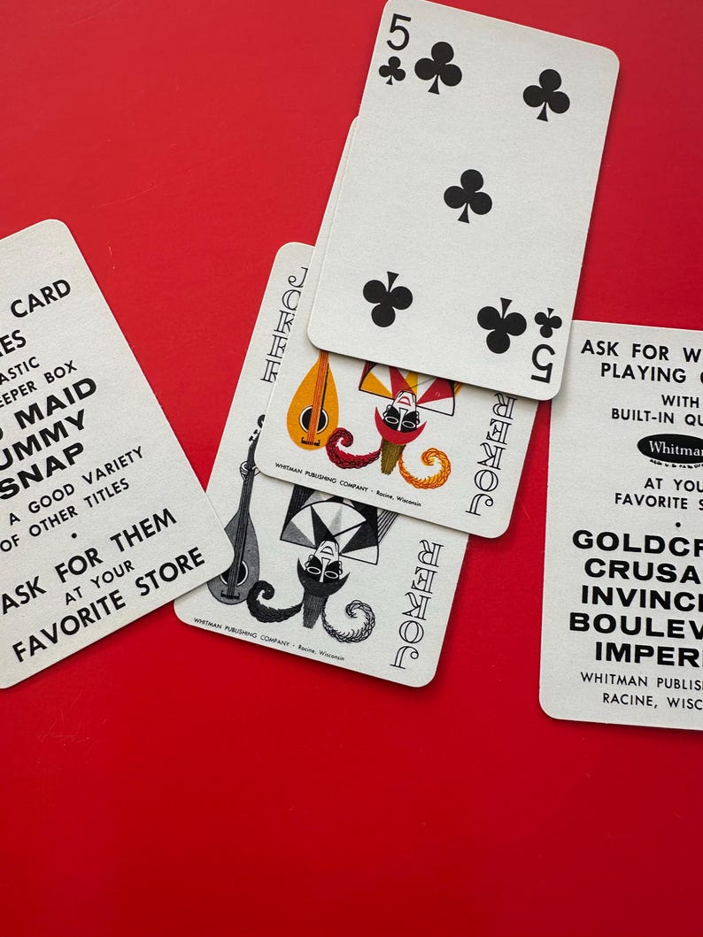 May include: A collection of vintage playing cards, including a five of clubs and two joker cards, arranged on a vibrant red surface. The cards feature classic designs and text from the Whitman Publishing Company.