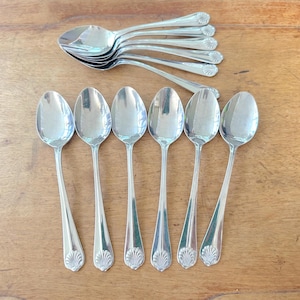 Może przedstawiać: Zestaw srebrnych łyżek z dekoracyjnym wzorem muszli na uchwytach. Łyżki ułożone są na drewnianej powierzchni, niektóre są ułożone w stos. Łyżki są odblaskowe i wydają się być wykonane ze stali nierdzewnej.