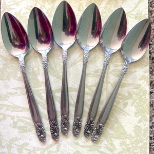 May include: Six silver spoons with floral detailing on the handles are arranged on a light green patterned surface. The spoons have a reflective surface, and a stainless steel serving utensil is at the top.