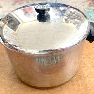 May include: A stainless steel cooking pot with a polished finish, featuring two black side handles and a matching lid with a central black knob. The pot has a cylindrical body and a reflective surface that shows light reflections from the surrounding area.