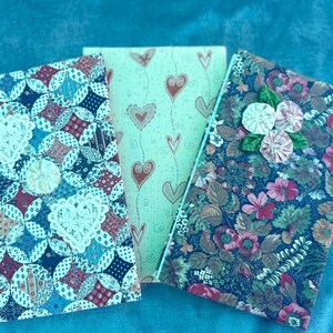 May include: Three fabric-covered journals with different patterns. The first journal has a blue and white patchwork design with a white lace heart. The second journal has a green background with a white heart pattern. The third journal has a dark blue background with a floral pattern and a white flower embellishment.