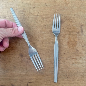Può includere: Due forchette d'argento su una superficie di legno. Una forchetta è tenuta in mano, mentre l'altra è piatta. Le forchette hanno un design semplice ed elegante con un manico sottile e quattro rebbi.