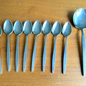 Könnte beinhalten: Ein Satz von acht silbernen Löffeln und einer silbernen Schöpfkelle. Die Löffel sind alle gleich groß und gleich geformt und haben ein schlichtes, modernes Design. Die Schöpfkelle ist größer als die Löffel und hat eine abgerundete Schüssel.
