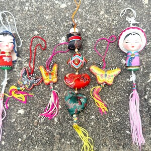 Pode incluir: Uma coleção de ornamentos coloridos de inspiração asiática. Os ornamentos incluem figuras de bonecas, formas de borboletas e corações, cada um com detalhes intrincados e borlas. A paleta de cores inclui vermelho, amarelo, rosa e verde.