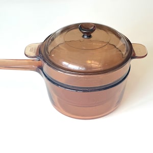 May include: A vintage amber-colored glass saucepan with a matching lid. The saucepan has a long handle and two side handles. The lid has a small knob. The cookware is made of transparent glass.