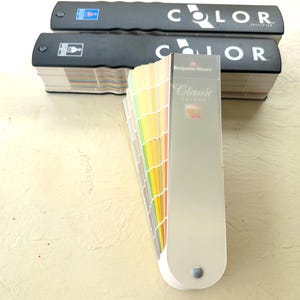 Op de afbeelding: Twee zwarte Benjamin Moore Color Specifier boeken horizontaal geplaatst achter een verticale Benjamin Moore Classic Colors kleurenwaaier. De waaier toont een breed scala aan kleurstalen in een spectrum, voorzien van een doorzichtige plastic hoes.