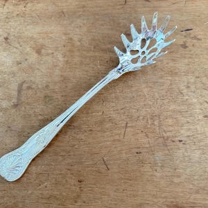 May include: A silver-toned metal pasta server featuring a long handle with an embossed shell-like pattern at the base and a wide, pronged head with a central cutout design. The utensil has a polished, reflective finish and a slender, tapered neck.