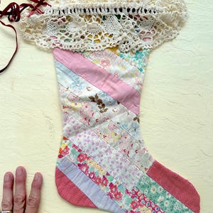 May include: A quilted Christmas stocking featuring a diagonal patchwork design of various floral and solid-colored fabrics in pink, green, and white. The top is trimmed with a wide, off-white crocheted lace cuff and includes thin dark red ribbons.