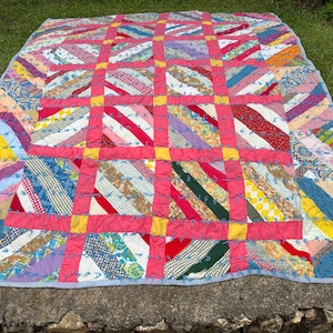 May include: A colorful handmade quilt with a patchwork design. The quilt features a grid pattern with pink, yellow, and red accents. Diagonal stripes in various colors and patterns fill the squares, creating a vibrant and cozy textile.