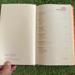 May include: Open cookbook with a red patterned cover, displaying the table of contents. Sections include appetizers, main fare, side courses, and special dishes. The book is open to pages with text and a copyright notice.