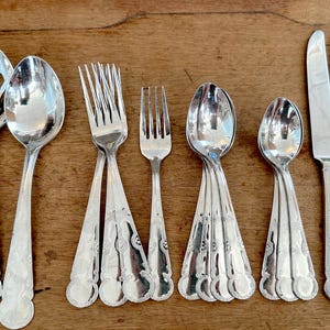 May include: An assortment of silver-colored flatware, including forks, spoons, and knives, arranged on a wooden surface. The utensils have decorative handles and are designed for dining. The arrangement includes various sizes and styles of forks, spoons, and knives.
