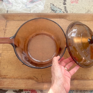 May include: Amber-colored glass saucepan with a long handle and a matching lid. The lid has a dark brown knob. The saucepan is sitting on a wooden cutting board.