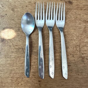 May include: A collection of vintage cutlery, comprising three forks and a spoon, displayed on a wooden surface. The silverware is silver-coloured, featuring a starburst design on the handles. The spoon is positioned on the left, with the forks to the right.