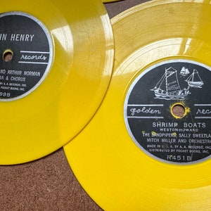 May include: Two yellow vinyl records with black labels. The top record has the text "JOHN HENRY golden records" and "WIN STRACKE AND ARTHUR NORMAN ORCHESTRA & CHORUS" with the record number "R499B". The bottom record has the text "golden records SHRIMP BOATS WESTON-HOWARD THE SANDPIPERS, SALLY SWEETLAND MITCH MILLER AND ORCHESTRA" with the record number "R451B".