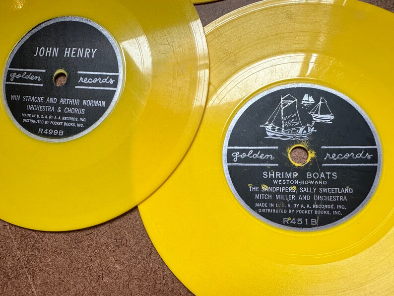 May include: Two yellow vinyl records with black labels. The top record has the text "JOHN HENRY golden records" and "WIN STRACKE AND ARTHUR NORMAN ORCHESTRA & CHORUS" with the record number "R499B". The bottom record has the text "golden records SHRIMP BOATS WESTON-HOWARD THE SANDPIPERS, SALLY SWEETLAND MITCH MILLER AND ORCHESTRA" with the record number "R451B".