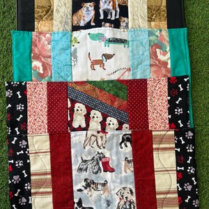May include: A patchwork dog bed featuring various dog breeds. The top section has bulldog images on a black background. The middle section has dachshunds and other dogs. The bottom section has various dog breeds and paw prints. The bed has a variety of colors.