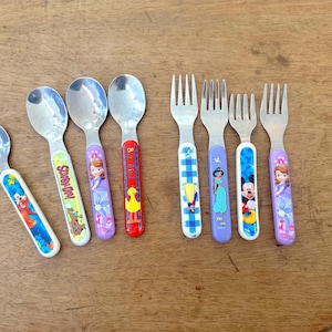 May include: A collection of children's silverware, including spoons and forks. The handles feature colorful cartoon characters such as Scooby-Doo, Sofia the First, and Mickey Mouse. The spoons and forks are made of stainless steel.