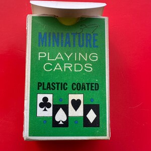 May include: A green box of miniature playing cards with the words "MINIATURE PLAYING CARDS PLASTIC COATED" in white and blue. The box features the suits of clubs, spades, hearts, and diamonds. The background is red.