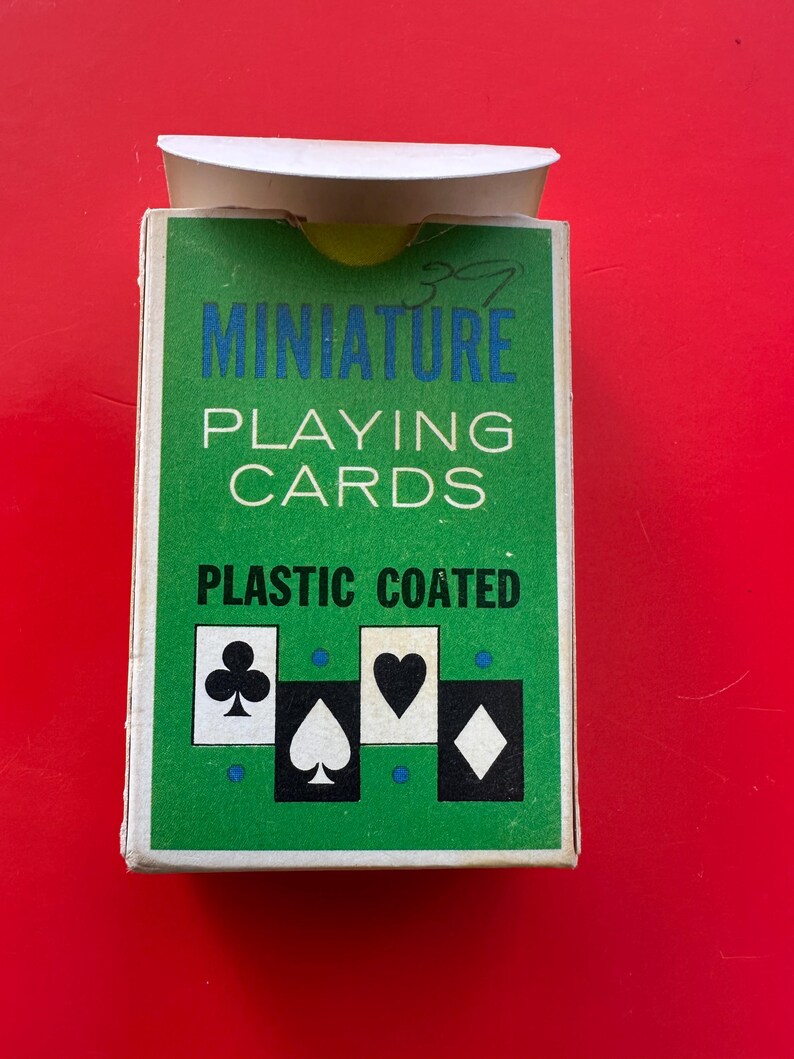 May include: A green box of miniature playing cards with the words "MINIATURE PLAYING CARDS PLASTIC COATED" in white and blue. The box features the suits of clubs, spades, hearts, and diamonds. The background is red.