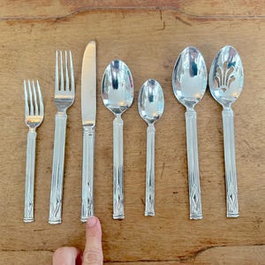 May include: A collection of silver-colored flatware, including forks, knives, and spoons, arranged on a wooden surface. The spoons vary in size, with one featuring a decorative openwork design. The handles have a vertical ribbed pattern.