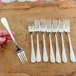 May include: A set of seven silver-toned metal dinner forks with a smooth, polished finish. Each fork features a slender handle that widens slightly toward the base and four tines. The forks are arranged in a row with one held separately.