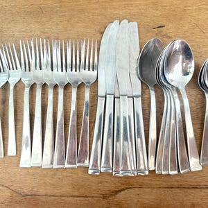 May include: A collection of stainless steel silverware, including forks, knives, and spoons, arranged on a wooden surface. The silverware has a simple, modern design with a polished finish. The forks have four tines, and the knives have a straight blade.