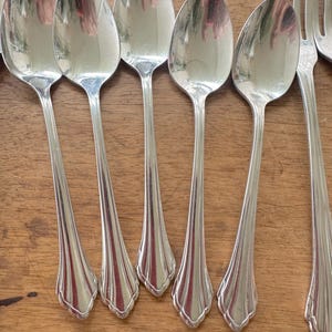 May include: A collection of silver spoons and forks arranged on a wooden surface. The spoons have a classic design with a teardrop-shaped bowl and a decorative handle. The silverware reflects light, creating a shiny appearance.
