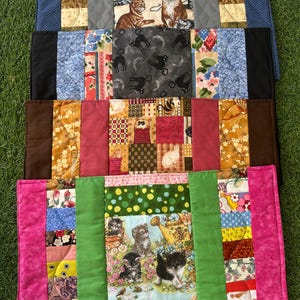 May include: A stack of colorful quilted placemats with cat and kitten patterns. The placemats feature various fabrics, including floral, polka dot, and cat-themed prints. The edges are trimmed with solid colors like pink, green, and black.