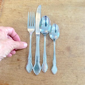 May include: Silverware set including a fork, knife, and two spoons. The utensils have decorative handles and are arranged on a wooden surface. The fork has four tines, and the knife has a smooth blade.