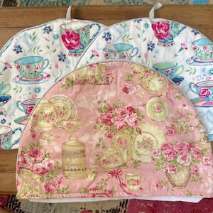 May include: Three tea cosies with floral patterns. Two are white with teacups and floral designs in pink, blue, and green. The third is pink with roses, teacups, and teapots. Each cosy has a loop for hanging.
