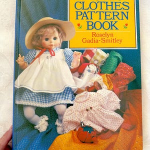 Könnte beinhalten: Ein Buch mit dem Titel "Dolls' Clothes Pattern Book" von Roselyn Gadia-Smitley. Der Umschlag zeigt eine Puppe, die ein blau-weiß kariertes Kleid und einen Strohhut trägt. Die Puppe ist umgeben von Stoffresten, Nähzubehör und einem kleinen weißen Kleid.