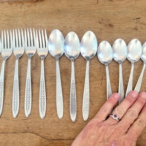 May include: A collection of vintage silverware, including forks and spoons, arranged on a wooden surface. The silverware has a shiny, silver finish with decorative detailing on the handles. The image showcases a set of dining utensils.