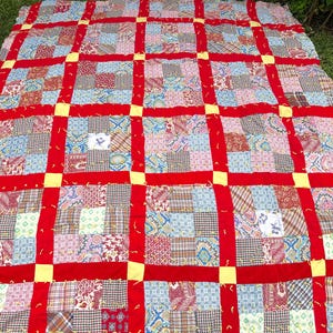 May include: A patchwork quilt with a grid pattern. The quilt features squares of various patterned fabrics in shades of blue, red, pink, and brown. Wide red bands form the grid, with yellow squares at the intersections. The quilt is laid out on green grass.