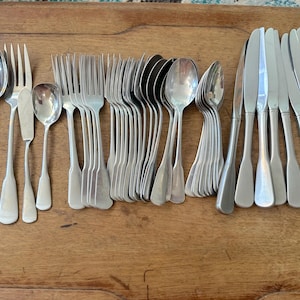 May include: A selection of stainless steel cutlery, including spoons, forks, knives, and butter knives, displayed on a wooden surface. The cutlery features a simple, classic design with rounded handles and a polished finish. The set includes various sizes of spoons and forks.