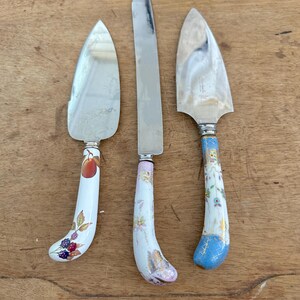 May include: A set of three serving utensils with silver-toned metal blades and curved ceramic handles. The left utensil features a peach and blackberry motif, the centre has a floral pattern, and the right has a blue and gold decorative design.