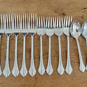 May include: A set of twelve silver-toned metal forks and four silver-toned metal spoons arranged in a row. Each piece of flatware features a slender handle with a flared, scalloped base and a polished, reflective finish on the tines and bowls.