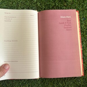 May include: Open cookbook with lined pages for recipes and a pink page with "Main Fare" and food categories: Beef, Lamb & Pork, Poultry, Seafood, and Stews. The book is open on a green surface.