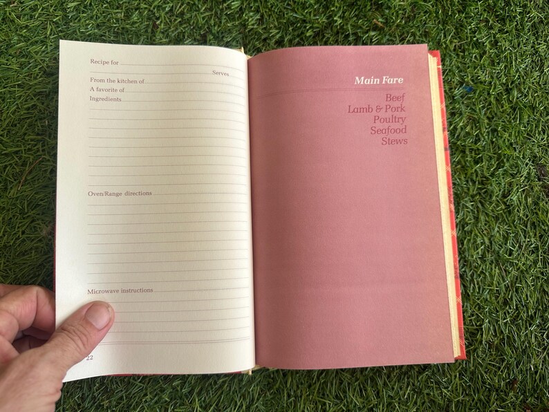 May include: Open cookbook with lined pages for recipes and a pink page with "Main Fare" and food categories: Beef, Lamb & Pork, Poultry, Seafood, and Stews. The book is open on a green surface.