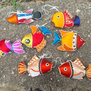Pode incluir: Uma coleção de ornamentos de peixes coloridos e feitos à mão. Cada peixe apresenta tons vibrantes de vermelho, laranja, amarelo e azul, com detalhes intrincados e acentos decorativos. São feitos de tecido e têm laços brancos para pendurar.