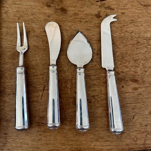 May include: A set of four silver-toned cheese serving utensils with smooth, tapered handles. The set includes a two-pronged fork, a rounded spreader, a triangular spade-shaped knife, and a narrow knife with a serrated edge and a forked tip.