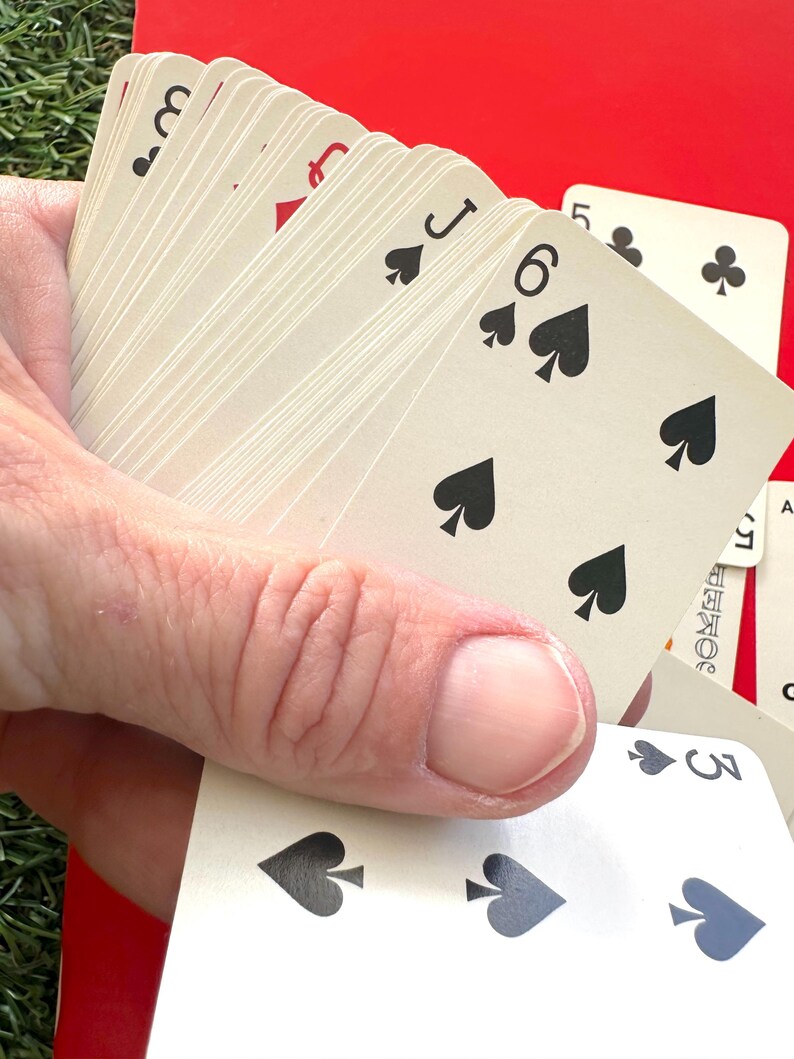 May include: A hand holding a fanned deck of playing cards. The cards are off-white with black and red suit symbols. Visible cards include the 3 of spades, 6 of spades, Jack of spades, and 5 of clubs. The background is red and green.